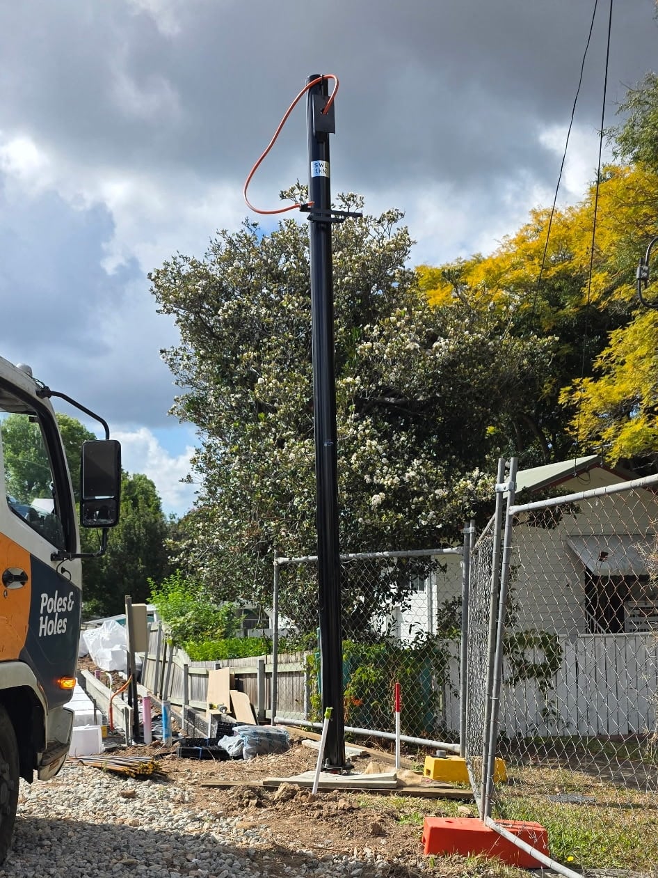 A black powder-coated permanent power pole standing on a residential building site, with the Poles and Holes truck parked alongside