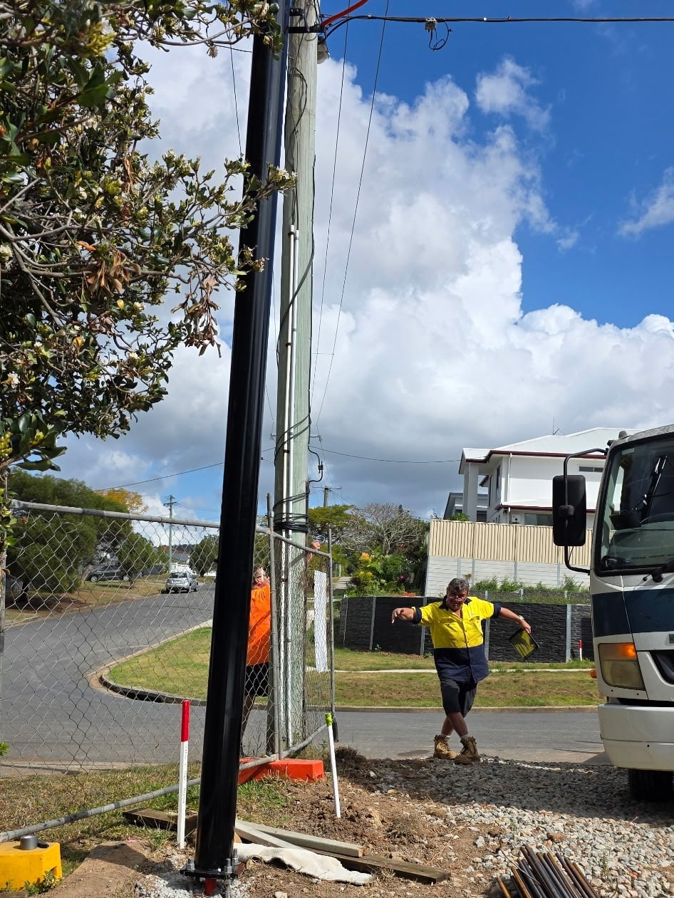 A black powder-coated permanent power pole bolted to its concrete footing, with the overhead service cable run down from the street pole and the crew on site