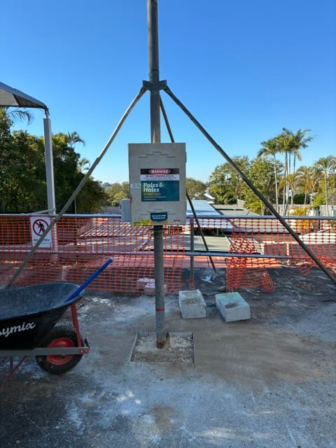 A stayed builder's power pole and meter box standing on a poured concrete slab behind safety fencing